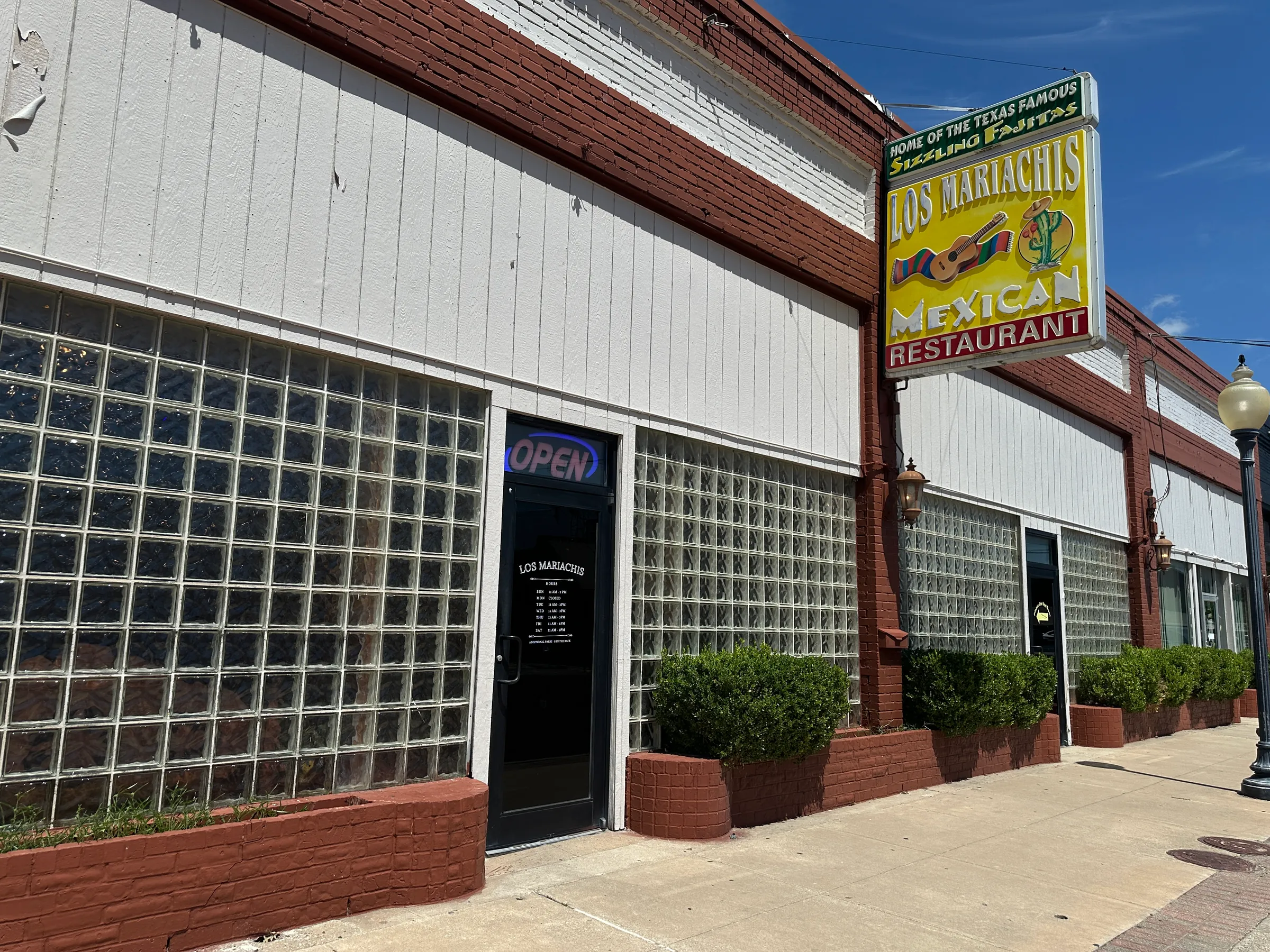 Sunny front entrance of Los Mariachis Mexican Restaurant with the bright yellow sign above the black door and glass block windows.